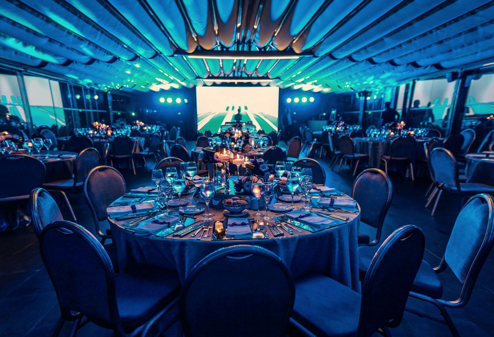 A ballroom set for a corporate gala dinner, with round tables and a stage screen